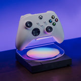 A white Xbox controller floating above a glowing iridescent charging pad on a dark stand, set against a purple-blue gradient studio background.